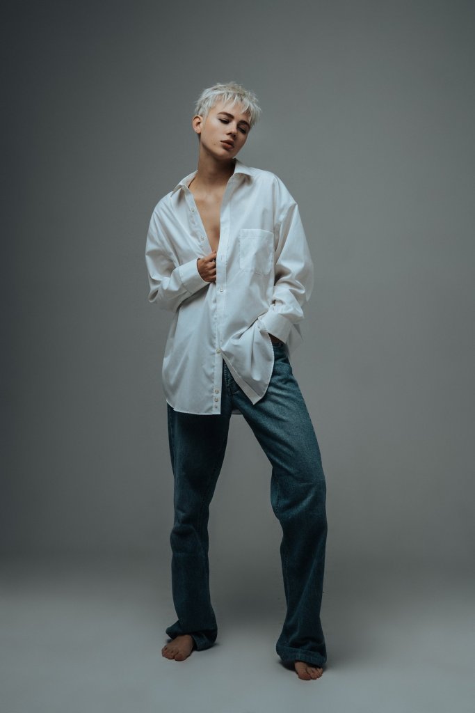 Model Chelsey Boll strikes a simple but effective pose in studio for a self portrait. She has her weight on one leg with the other bent, and one hand is in the pocket of her baggy jeans while the other suggestively but subtly reaches into her shirt. Her face is angled to the side, and her eyes are looking down.